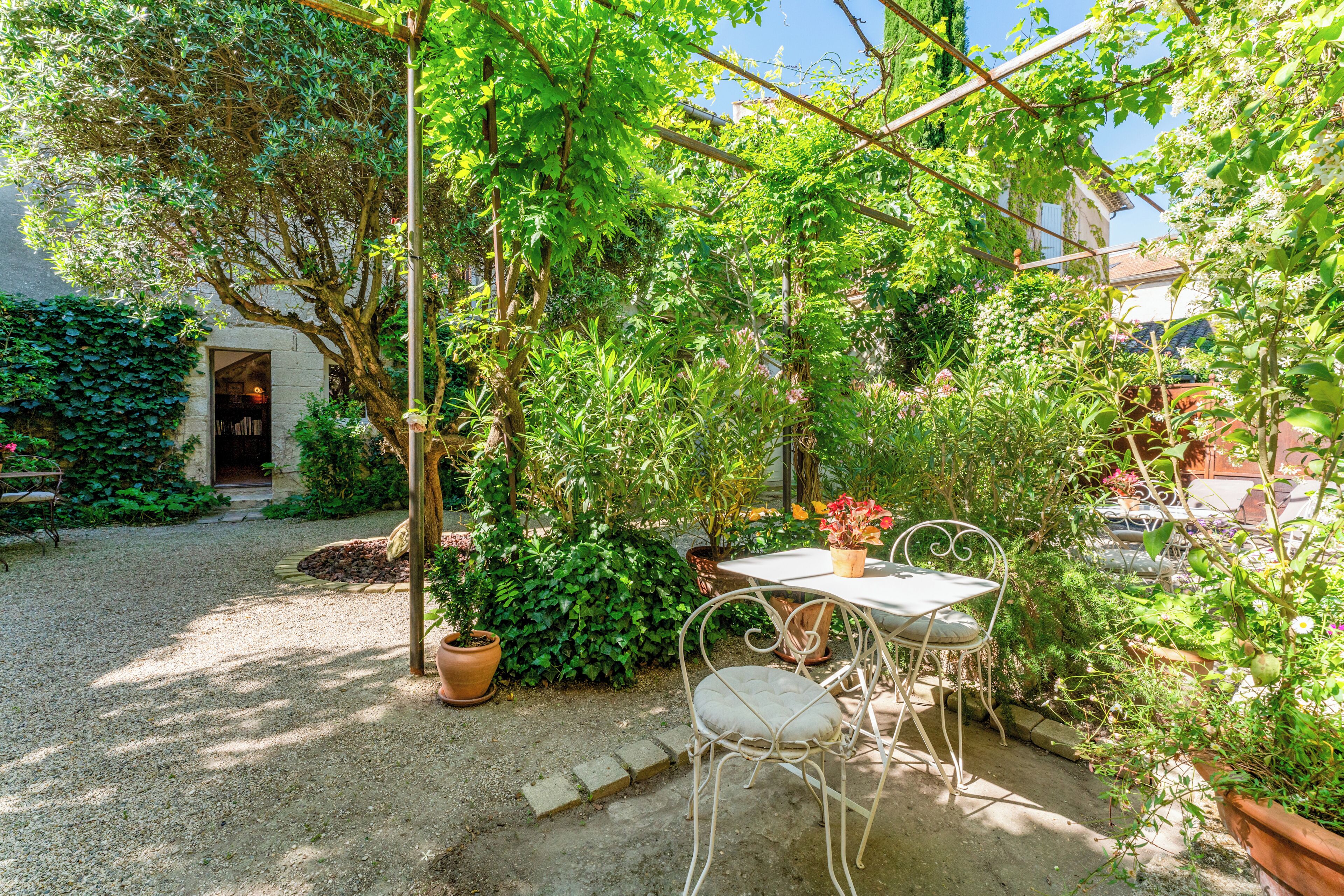 Eine Terrasse mit Tisch und Stühlen, umgeben von Grünpflanzen und einem Hauseingang.