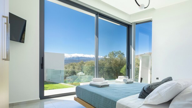 A modern bedroom with a large sliding glass door leading to a balcony, a bed with white and blue bedding, and a flat-screen TV mounted on the wall. - White View Villa by Alex – Your Dream Holiday in the Heart of Crete (Chania)