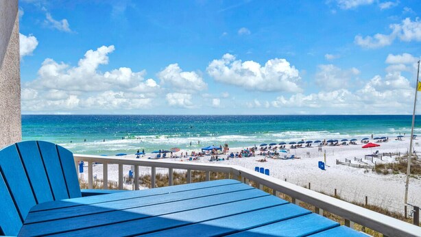 Une vue sur la plage depuis un balcon, avec des chaises bleues et une rambarde.