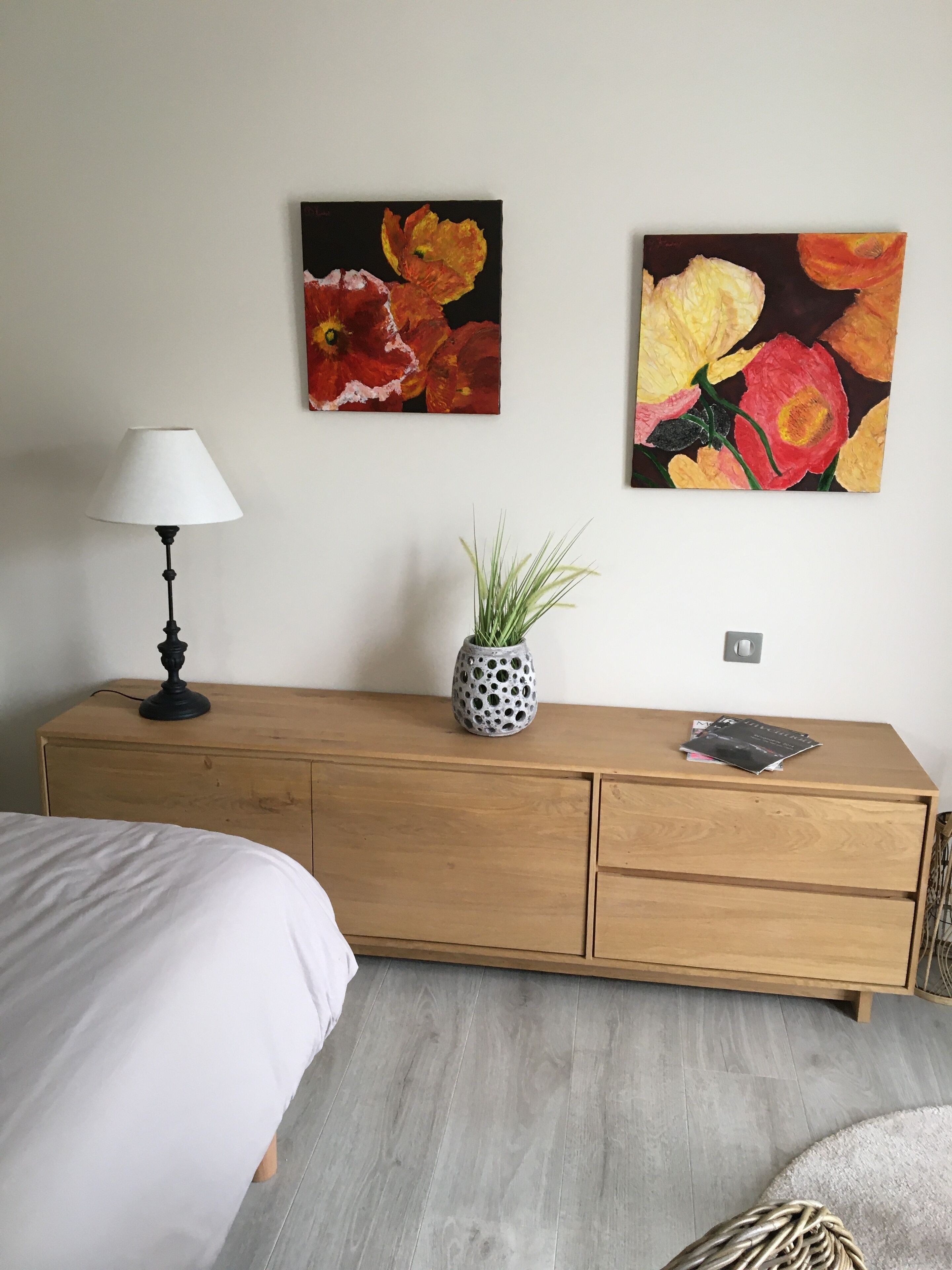 Un dormitorio con un aparador de madera, una cama, una lámpara y dos cuadros de flores en la pared.