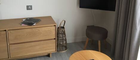 Ein modernes Wohnzimmer mit einem Holzschrank, einem runden Tisch mit Fernbedienung, einem wandmontierten Fernseher und einem Blumenbild.