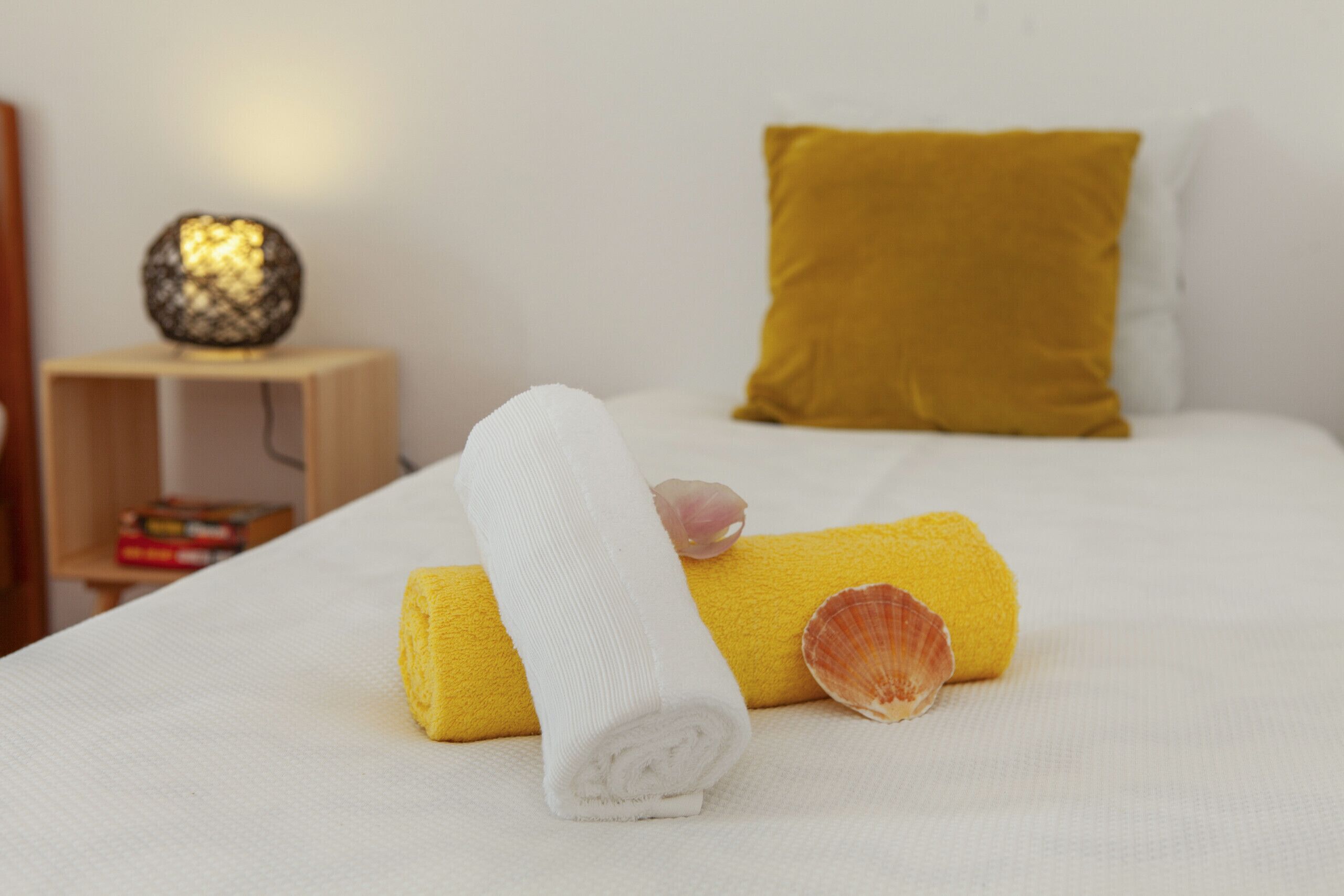 A neatly made bed with a yellow towel, a rolled white towel, and a seashell. There is a mustard-colored pillow and a small wooden nightstand with a decorative item and books.