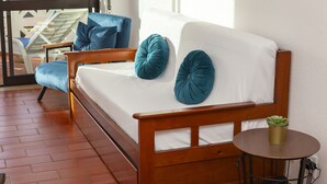 A wooden sofa with a white cushion and teal pillows, a small round table with a plant, and a view of a balcony with chairs.