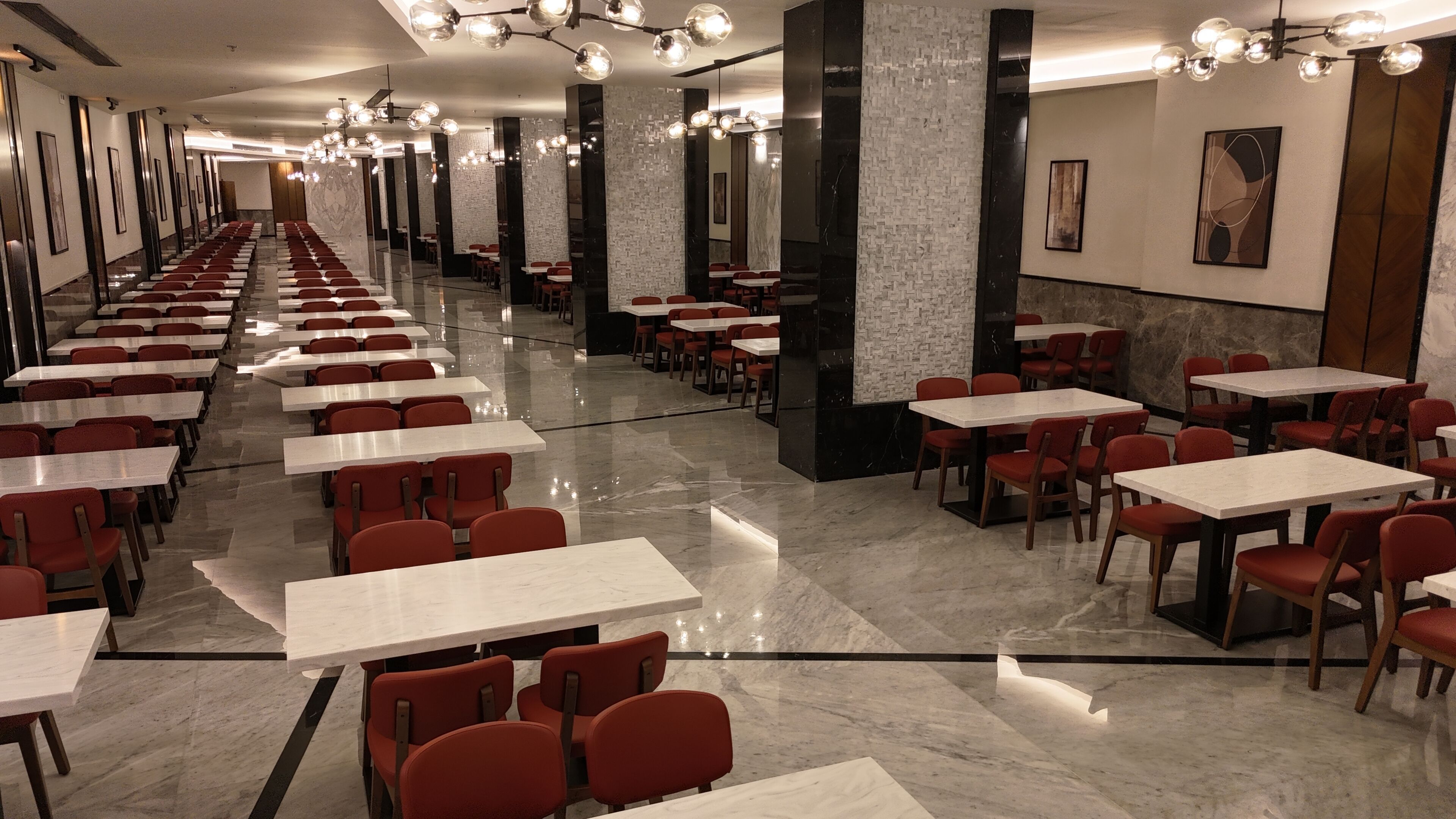 A spacious, well-lit dining area with long tables and red chairs, featuring a modern design with marble flooring and decorative lighting.