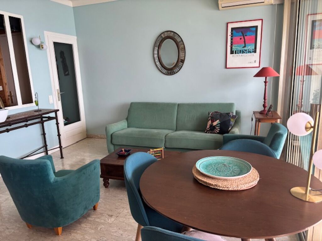 Una sala de estar con un sofá verde, sillones turquesa, una mesa de centro redonda de madera y una lámpara roja para el suelo.