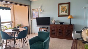 A living room with a teal armchair, a television, a cabinet, and a dining area with chairs and a table.