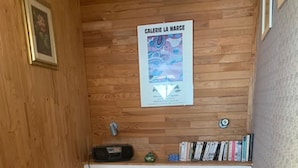 A small, cozy room with a wooden wall, a bed with a floral-patterned pillow, a shelf with books and a small radio, and a poster titled "GALERIE LA MARGE".