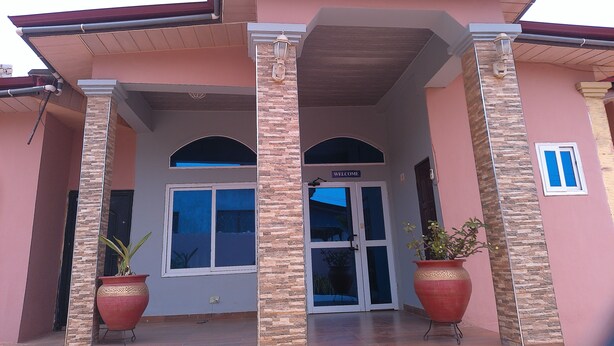 A building entrance with a 'WELCOME' sign, two large potted plants, and a tiled patio area. - Great Escape Inn Hotel  (Weija)
