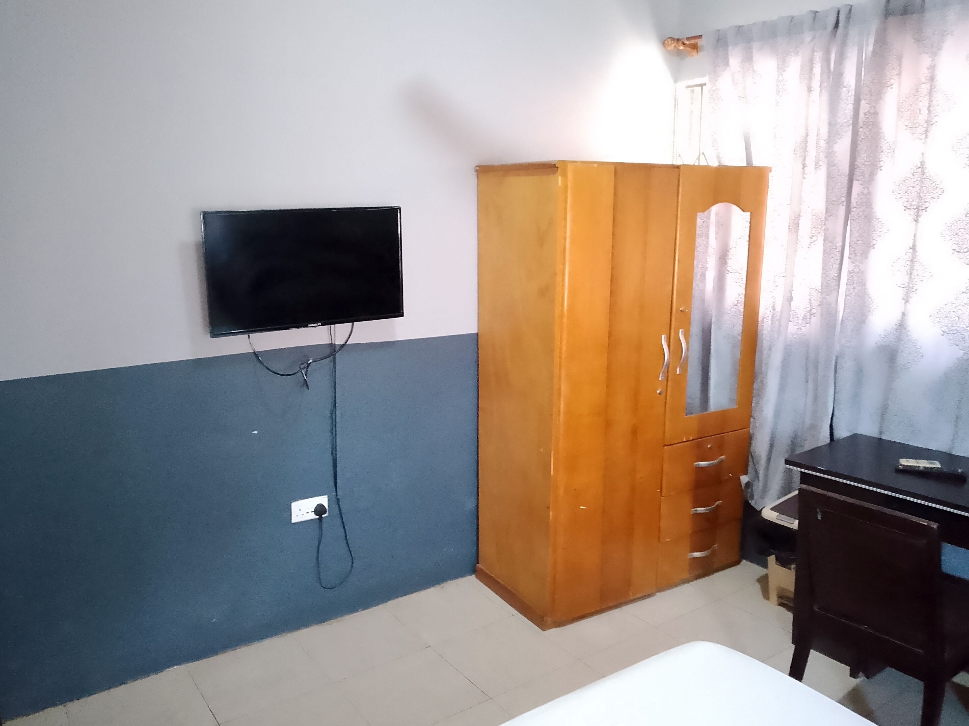 A room with a bed, a wardrobe, a desk, a television mounted on the wall, and a window with curtains.