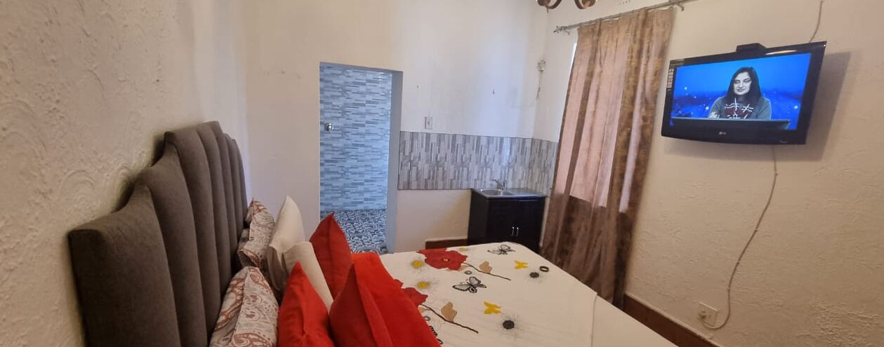 A hotel room with a bed, a television on the wall, a small table, and a window with curtains.