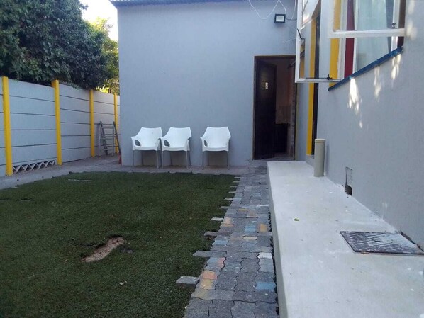 A small outdoor area with a grassy lawn, a stone pathway, and three white plastic chairs. - Neo & ruks guest house Boston (Cape Town)
