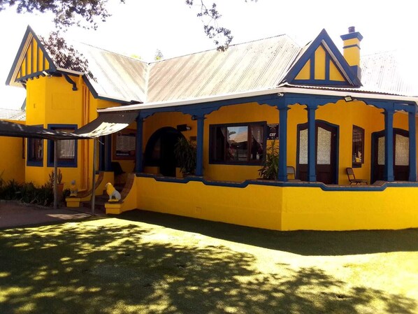 A yellow building with a blue roof and a chimney, featuring a porch with chairs and a small table. - Neo & ruks guest house Boston (Cape Town)