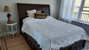 A neatly made bed with a white lace bedspread, a dark wooden headboard, and two pillows. There is a small bedside table with a lamp and a decorative horse figure. A window with curtains is visible, allowing natural light into the room. - Tranquil lakefront oasis - close to NYC and NJ attractions (Watchung)