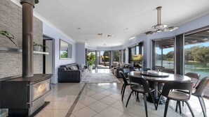 A modern living room with a fireplace, a dining area with a round table, and large windows offering a view of a lake. - Riverside Manor (Sussex Inlet)