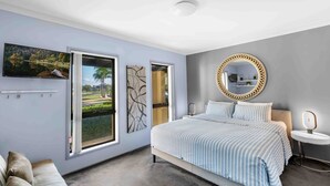 A modern bedroom with a large bed, a round mirror, a television, and a view of a residential area through the windows. - Riverside Manor (Sussex Inlet)