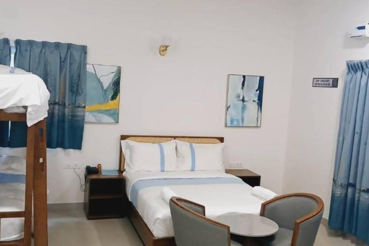 Ein Hotelzimmer mit Etagenbett, einem Einzelbett, einem Nachttisch, einem Stuhl und einem kleinen Tisch. An der Wand hängen zwei Bilder und ein Fenster mit Vorhängen.