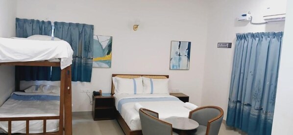 Ein Hotelzimmer mit Etagenbett, einem Einzelbett, einem Nachttisch, einem Stuhl und einem kleinen Tisch. An der Wand hängen zwei Bilder und ein Fenster mit Vorhängen.