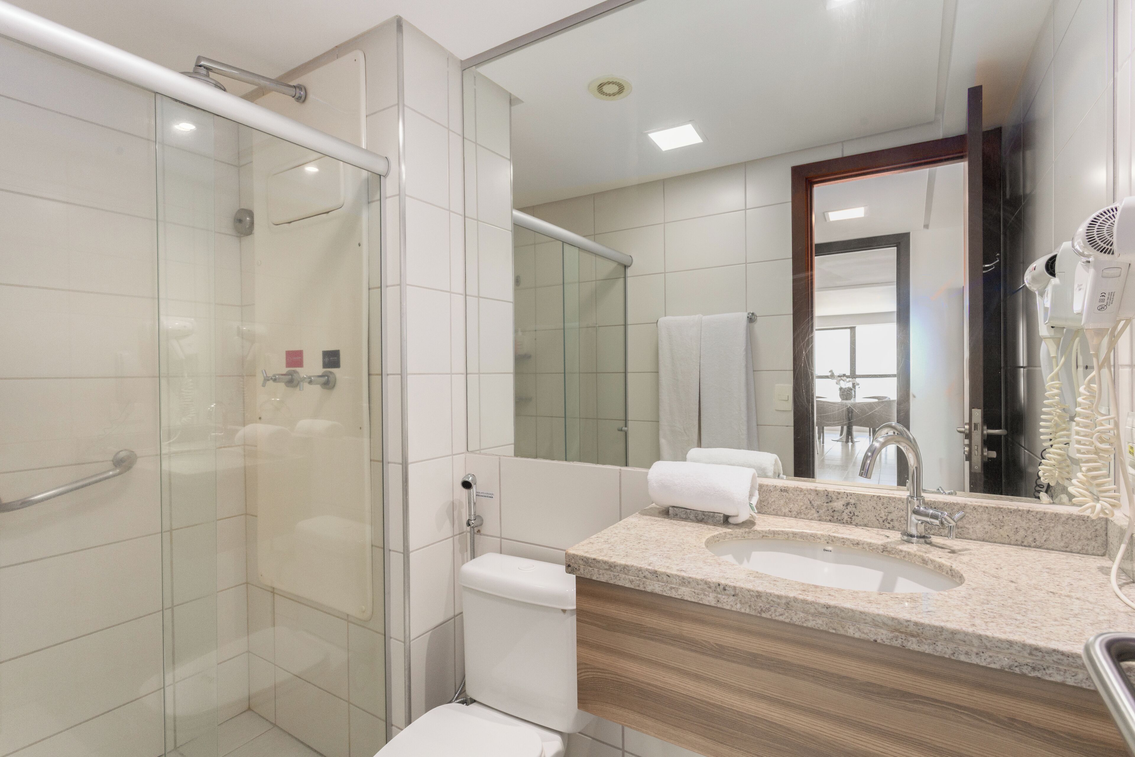 Un baño moderno con cabina de ducha de vidrio, inodoro, lavabo con mueble y un secador de pelo sobre un soporte.