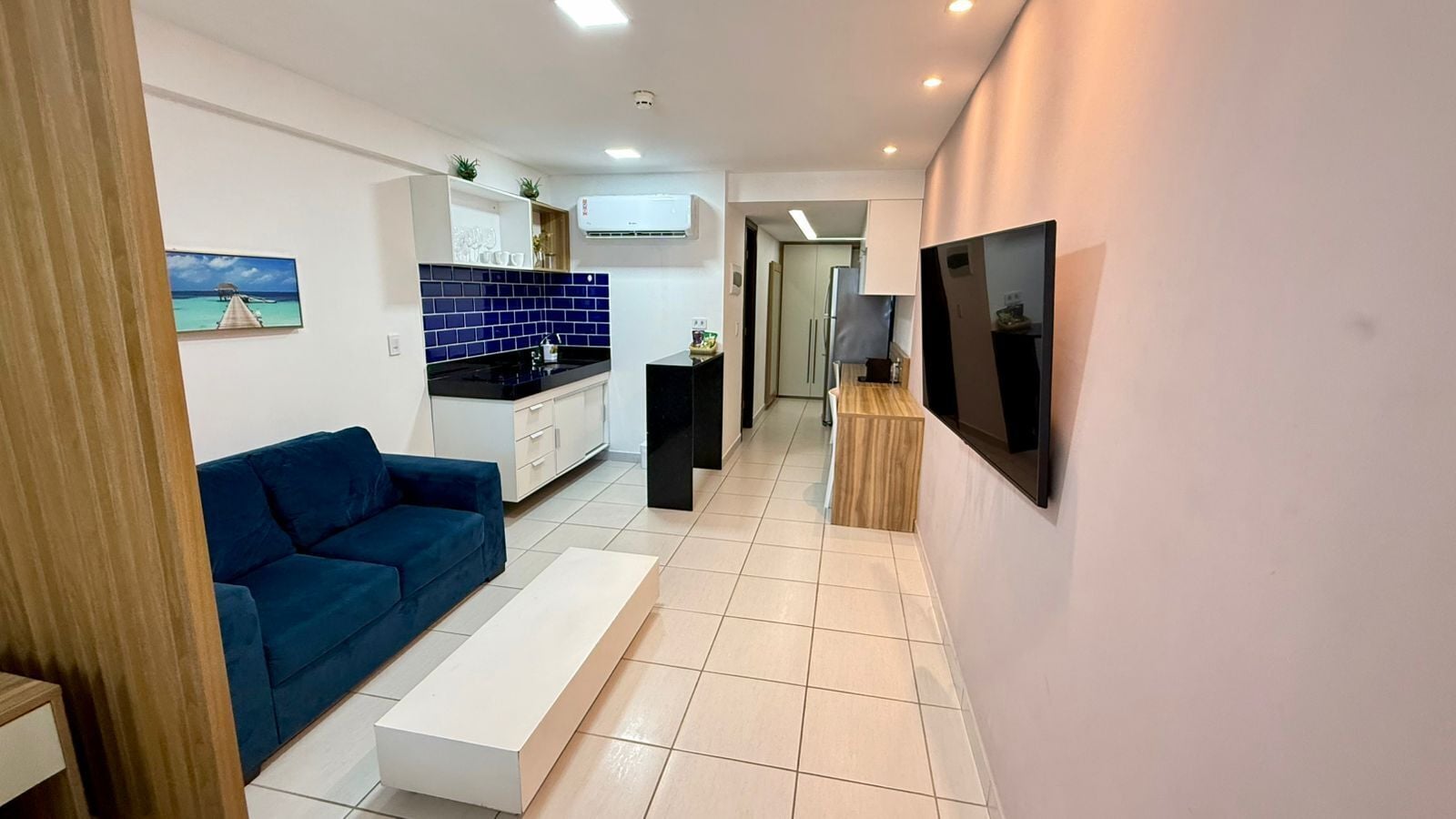Una sala moderna con un sofá azul, una mesa de centro blanca y una zona de cocina con azulejos azules y encimera negra.