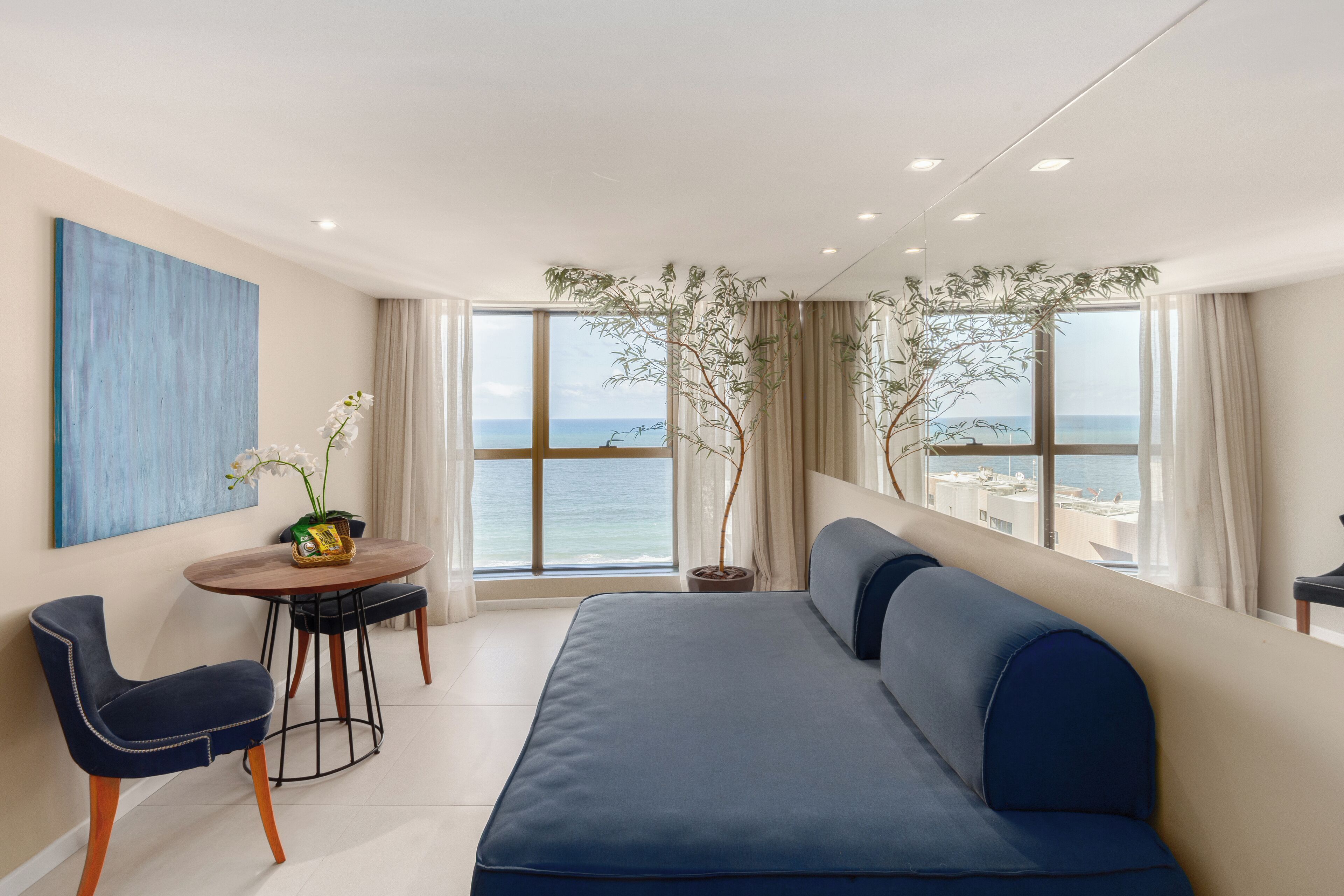 Ein modernes Zimmer mit großem Fenster und Meerblick, einem blauen Sofa, einem Esstisch mit Stühlen und einer Zimmerpflanze.