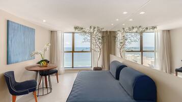Ein modernes Zimmer mit großem Fenster und Meerblick, einem blauen Sofa, einem Esstisch mit Stühlen und einer Zimmerpflanze.
