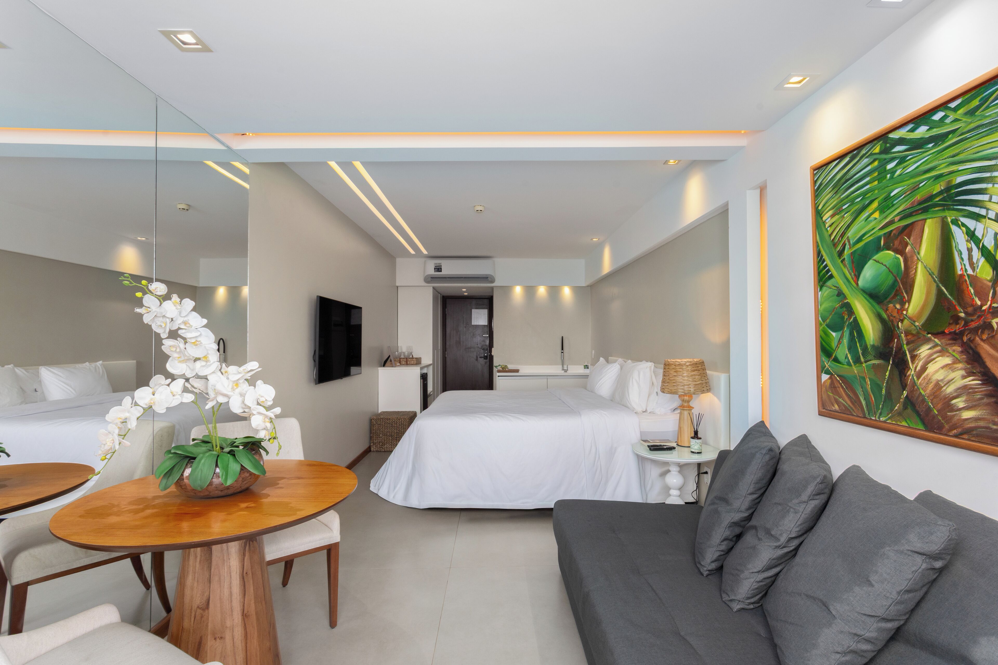 A modern hotel room with a bed, a sofa, a dining table with chairs, a small kitchen area, and a large window.