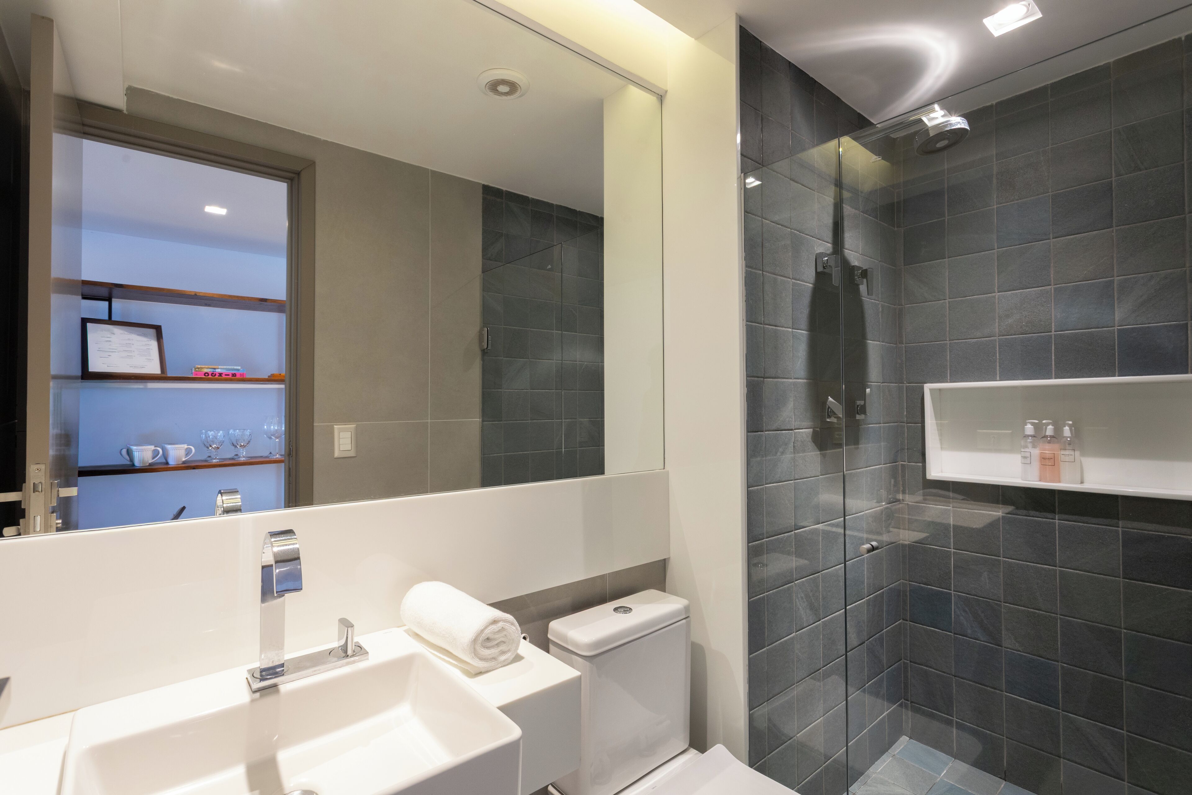 A modern bathroom with a white sink, a glass-enclosed shower, and a mirror reflecting a shelf with toiletries.