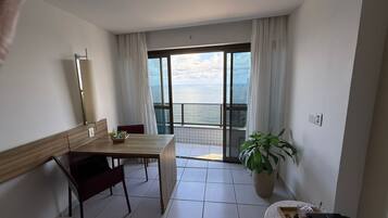 A room with a wooden table, chairs, a potted plant, and a view of the sea through large windows.
