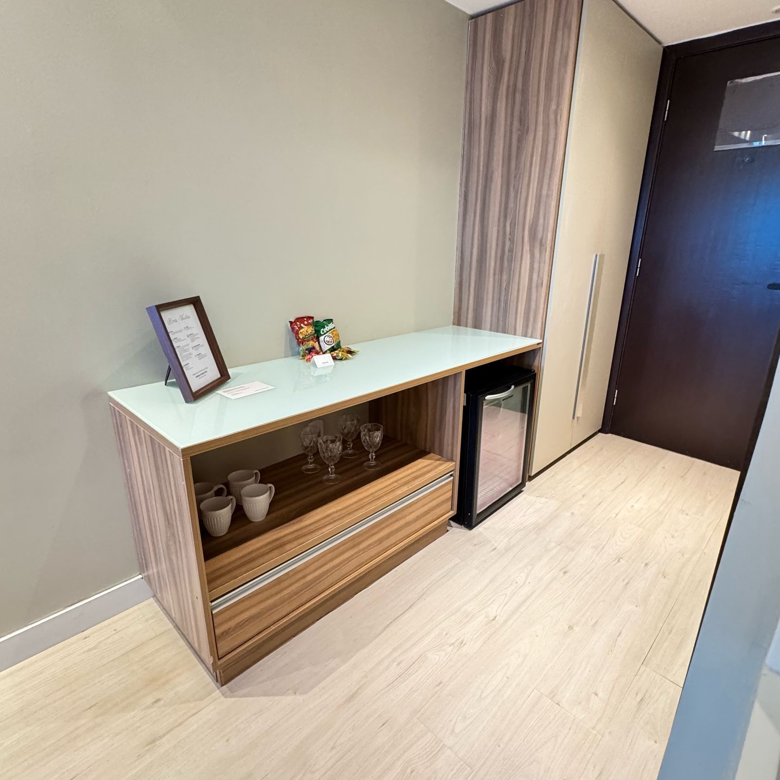 A small kitchenette with a light blue countertop, a wooden shelf with cups, and a refrigerator.
