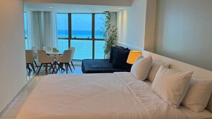 A hotel room with a large bed, a dining area with chairs, and a view of the sea.