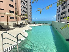 Una piscina con agua cristalina, rodeada de sillas de descanso y sombrillas, con vista al océano y un edificio de gran altura al fondo.