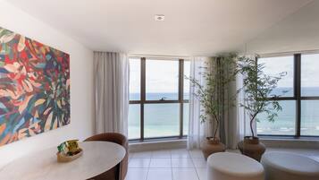 Una habitación moderna con un ventanal grande con vistas al mar, una mesa de comedor y una cesta de aperitivos, junto a dos macetas con plantas.