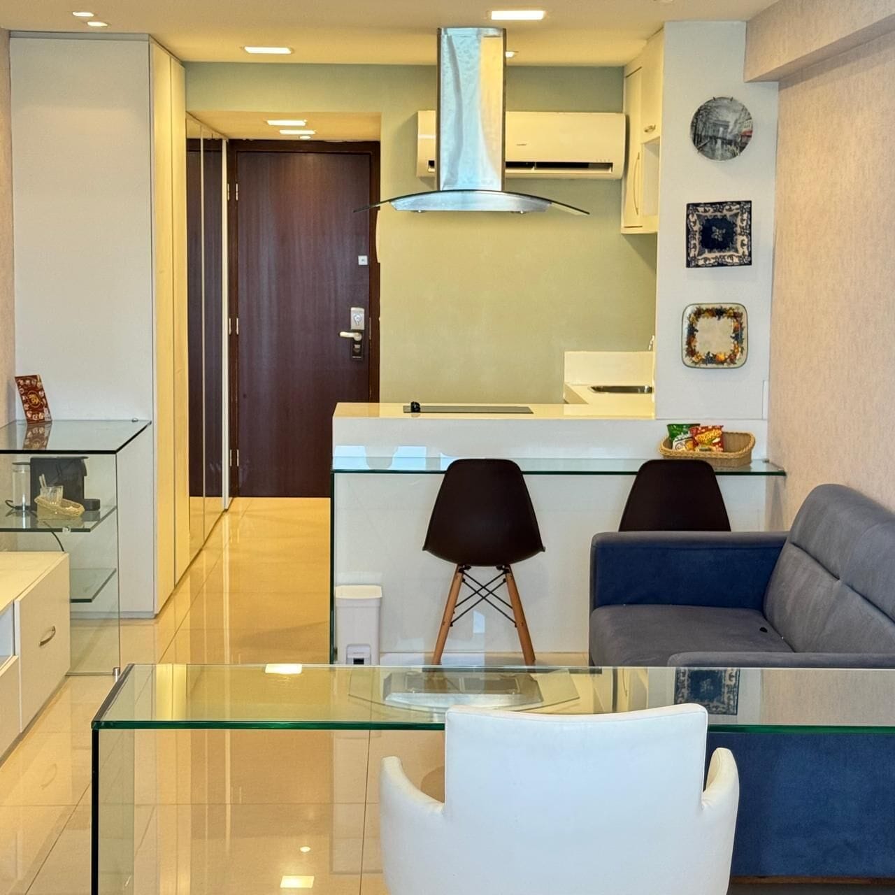 Una sala de estar moderna con una mesa de centro de vidrio, dos sillas negras, un sofá gris y una zona de cocina con encimera blanca y una campana extractora de acero inoxidable.
