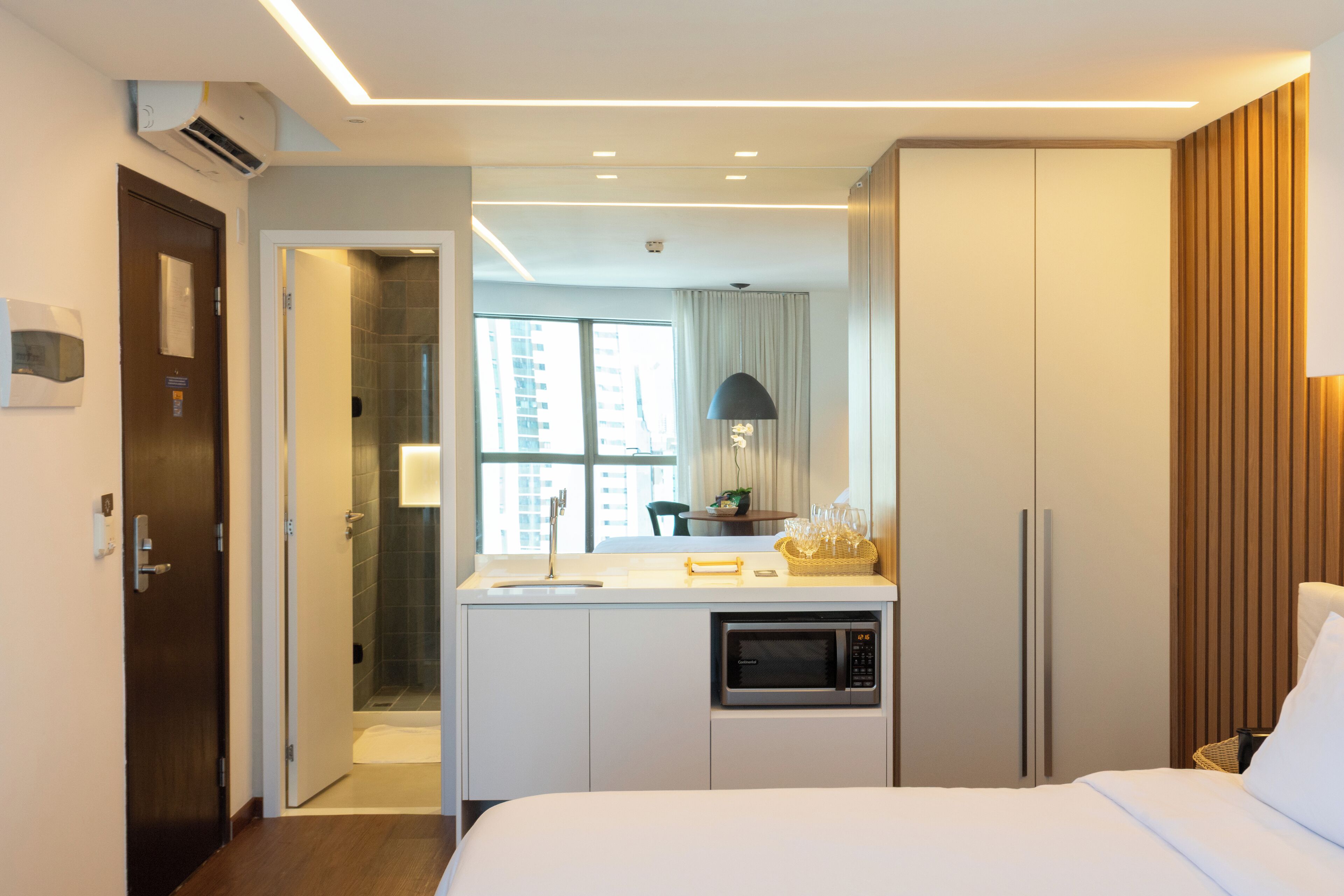 A hotel room with a bed, a kitchenette, a bathroom, and a view of the city.