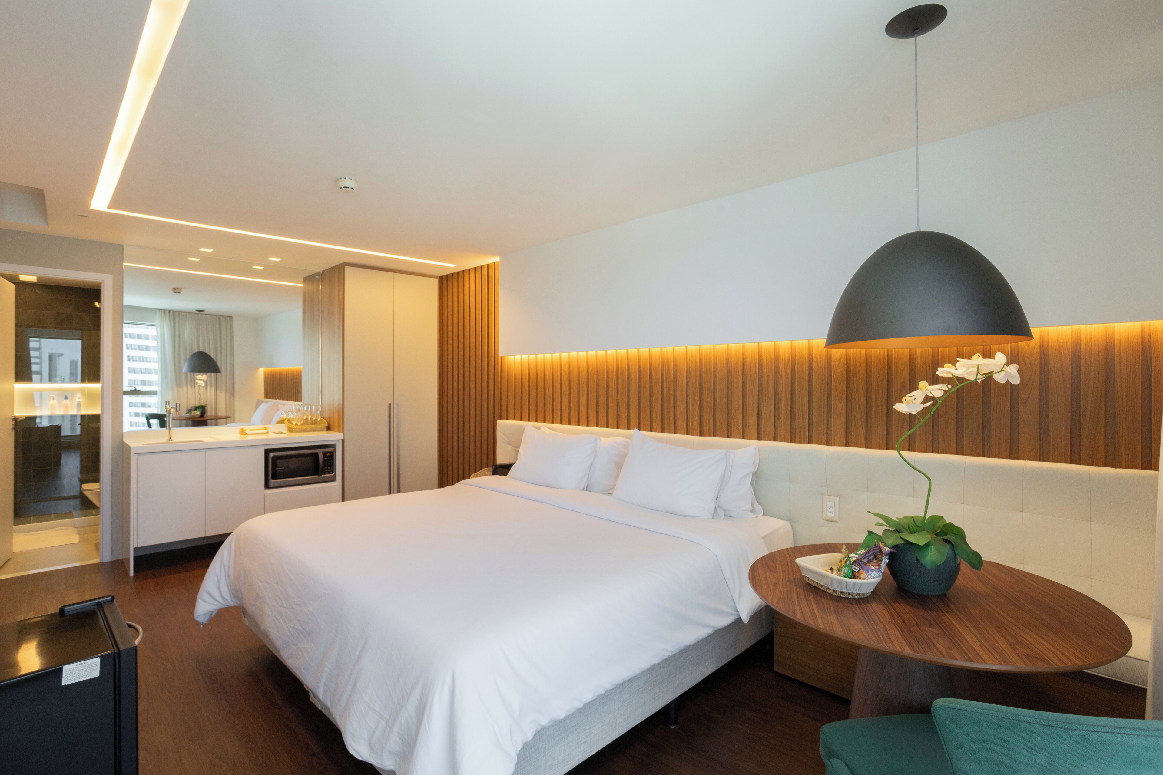 A modern hotel room with a large bed, a wooden headboard, a round wooden table with a potted plant, and a mirror above the bed.