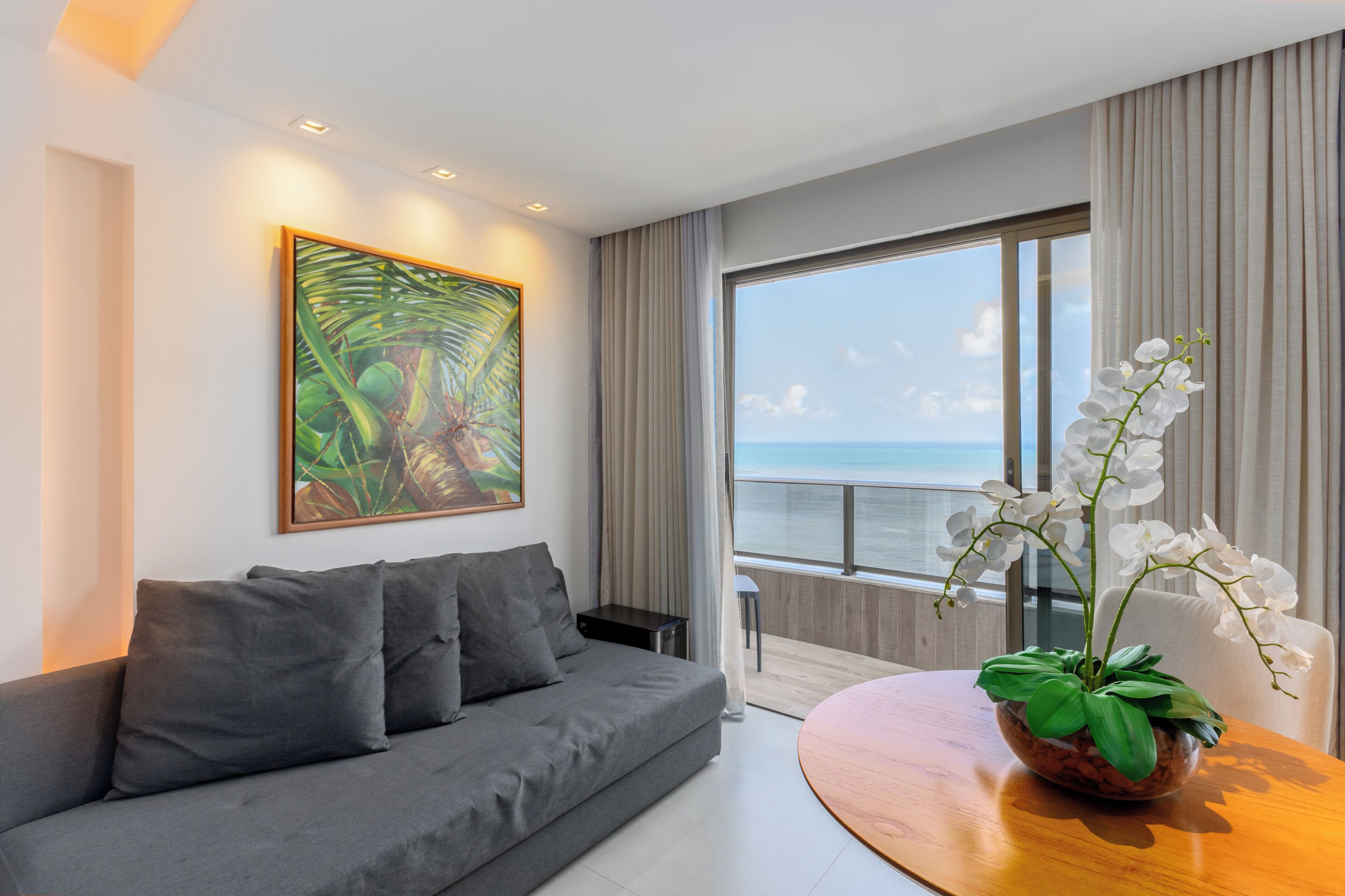 Un salon moderne avec un canapé gris, une table d’appoint en bois, une grande fenêtre donnant sur la mer et un tableau sur le thème des paysages tropicaux.