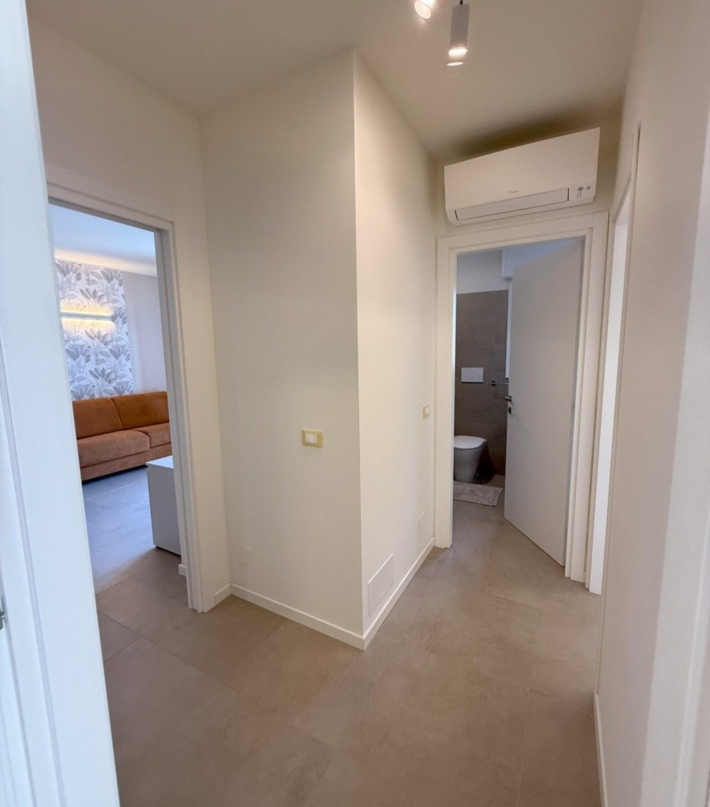 A hallway with a beige tiled floor, a white door leading to a bathroom, and a brown sofa in the background.