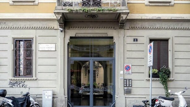 Entrada de edifício com porta de vidro, placa indicando "Via Giulio Urbati" e placa de proibido estacionar.