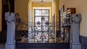 Um portão de ferro forjado ornamentado com desenhos intrincados, que dá acesso a um corredor com piso de mármore e uma grande porta em arco.