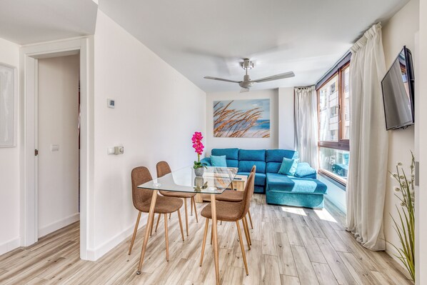A modern living room with a glass dining table, blue sofa, and a ceiling fan. - Apartamentos Donato Vista Mar by Croma (Estepona)