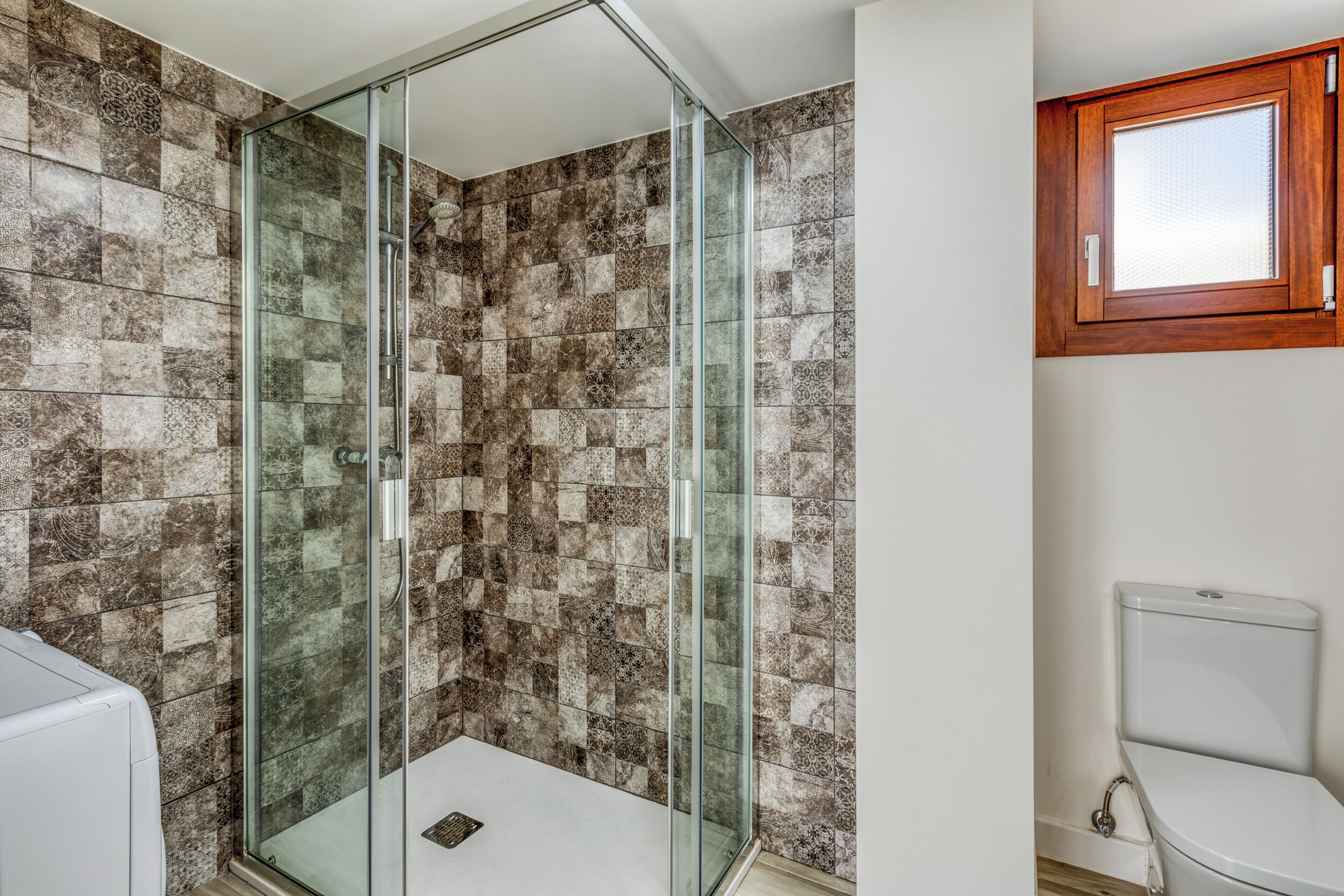 Une salle de bain avec une douche à parois de verre, des toilettes blanches et une fenêtre en bois.