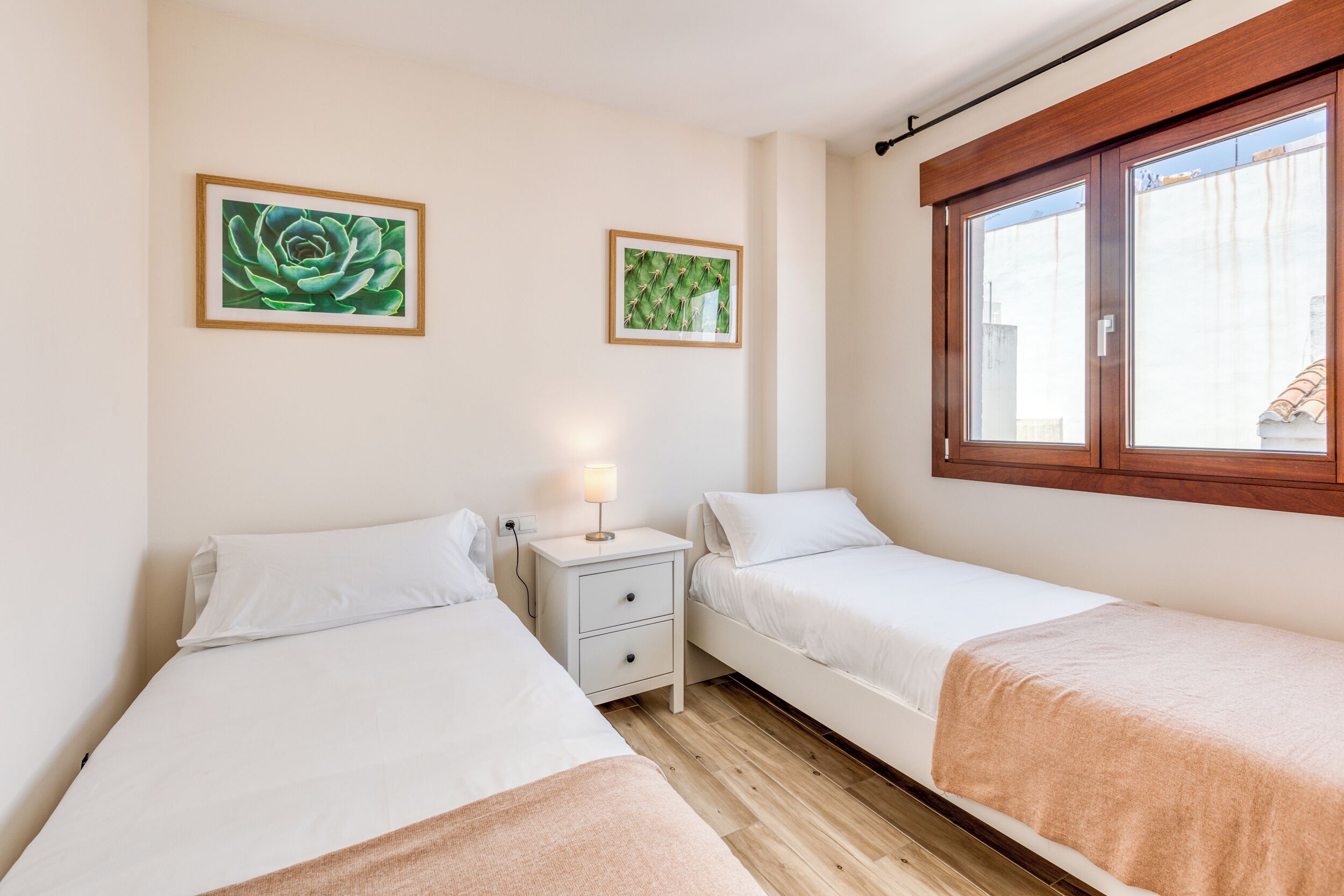 Un dormitorio con dos camas individuales, una mesita de noche con lámpara y dos cuadros enmarcados en la pared.