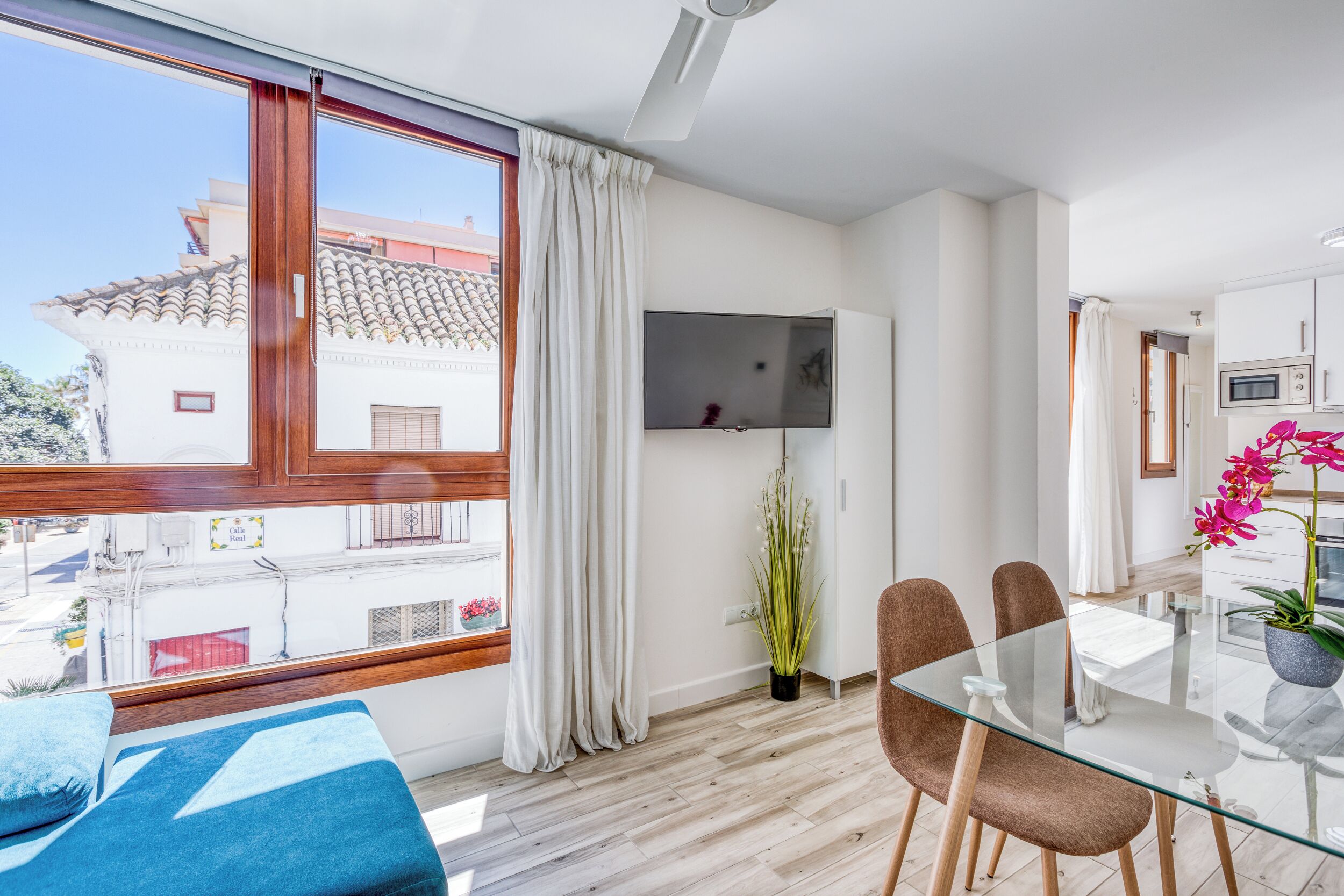 Ein modernes Wohnzimmer mit einem Glastisch, zwei braunen Sesseln, einem blauen Sofa, einem an der Wand befestigten Fernseher und einem großen Fenster mit weißen Vorhängen.