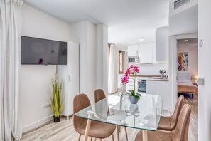 A modern living room with a glass dining table, brown chairs, a flat-screen TV mounted on the wall, a potted plant, and a view into the adjacent kitchen. - Apartamentos Donato Vista Mar by Croma (Estepona)