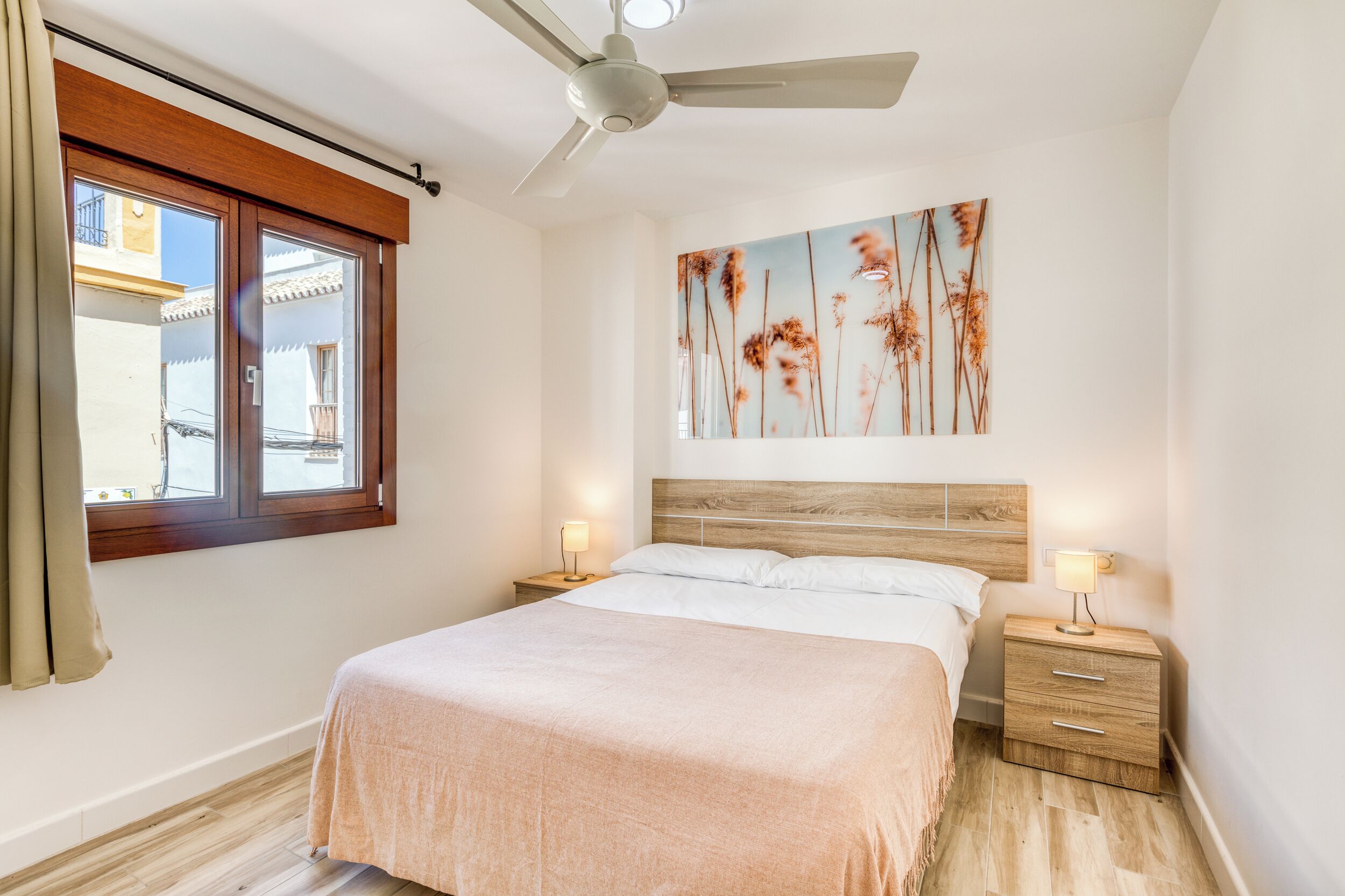 Un dormitorio con una cama, ventilador de techo, mesita de noche con lámpara, cabecera de madera y una obra de arte en la pared.