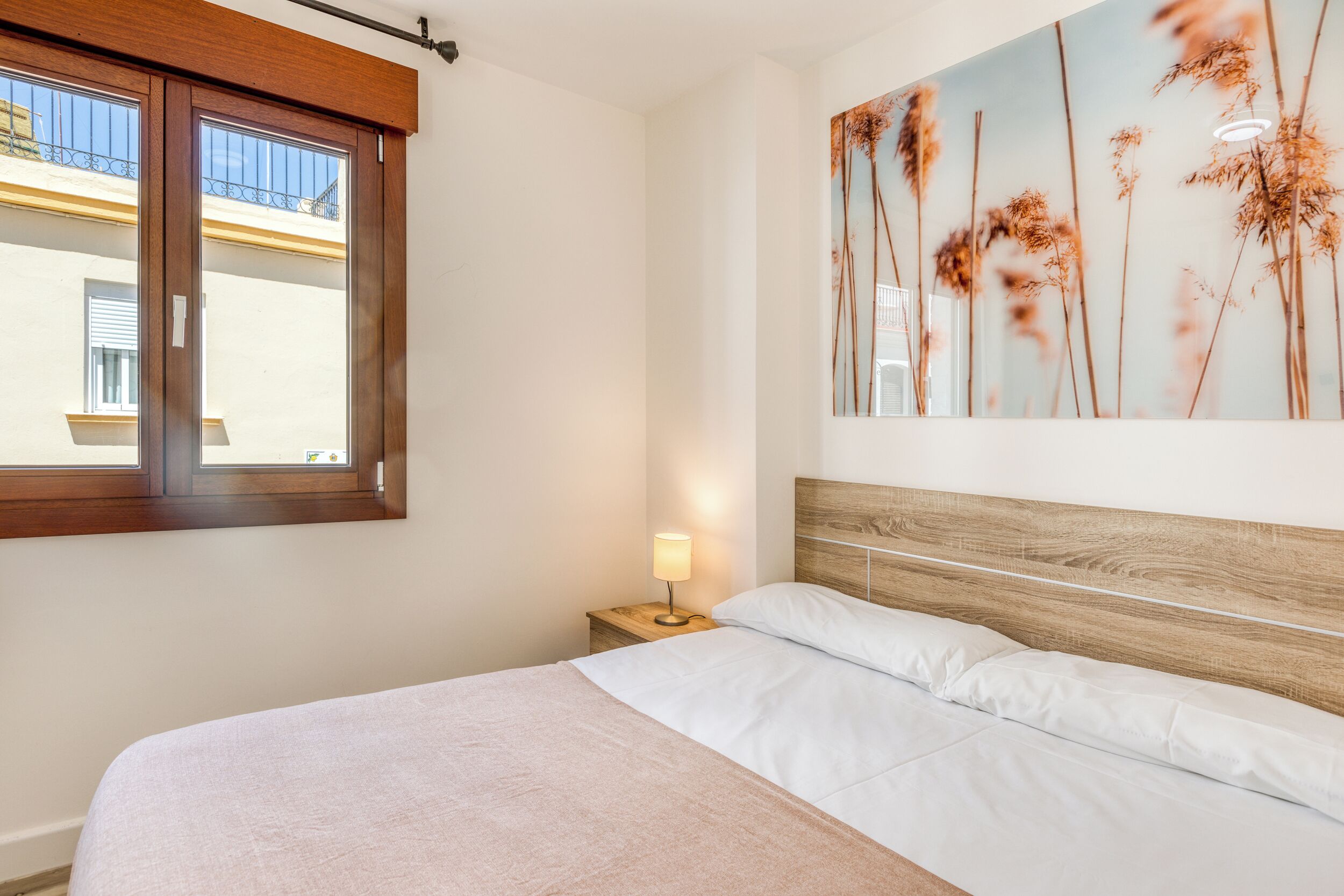 A neatly made bed with a wooden headboard, a bedside table with a lamp, and a window with blinds.