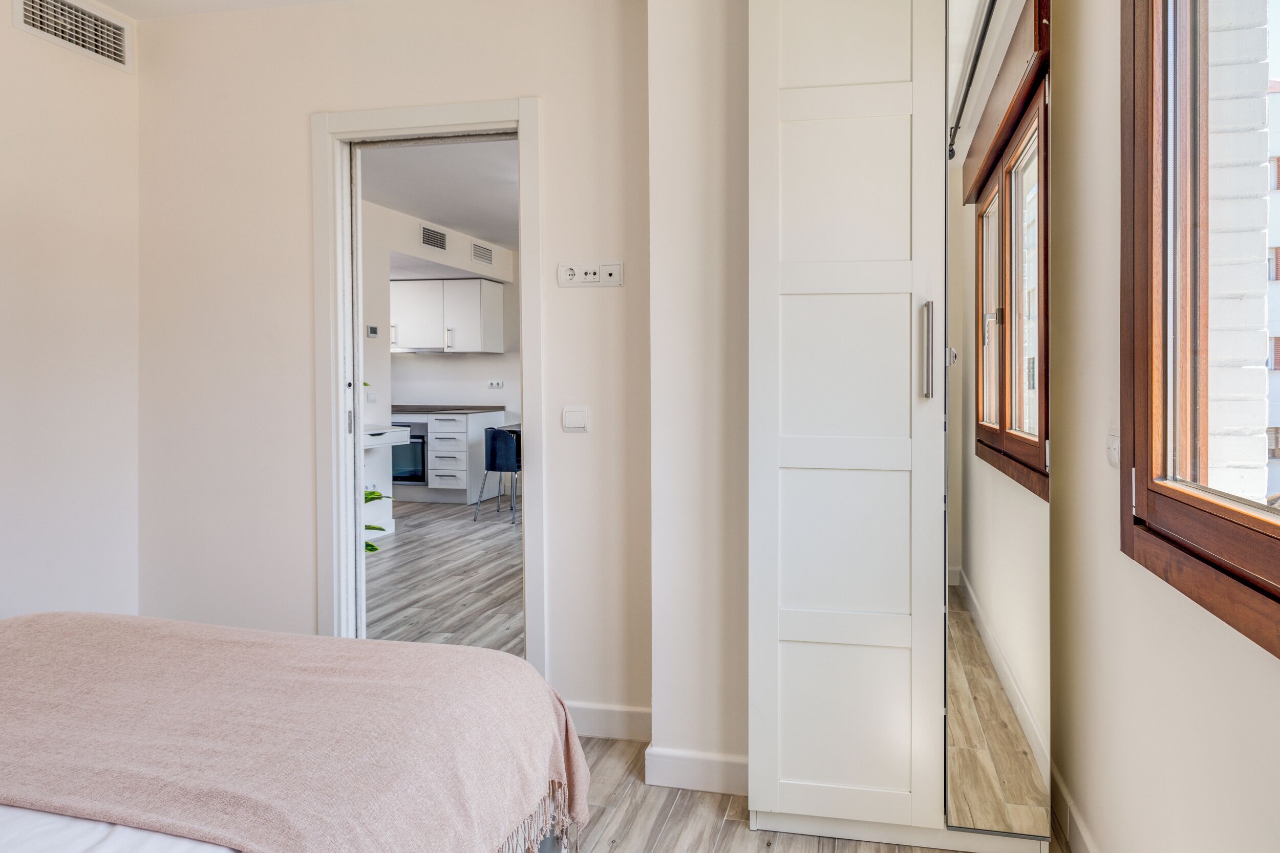 A bedroom with a bed, a door leading to a kitchen, and a window with blinds.