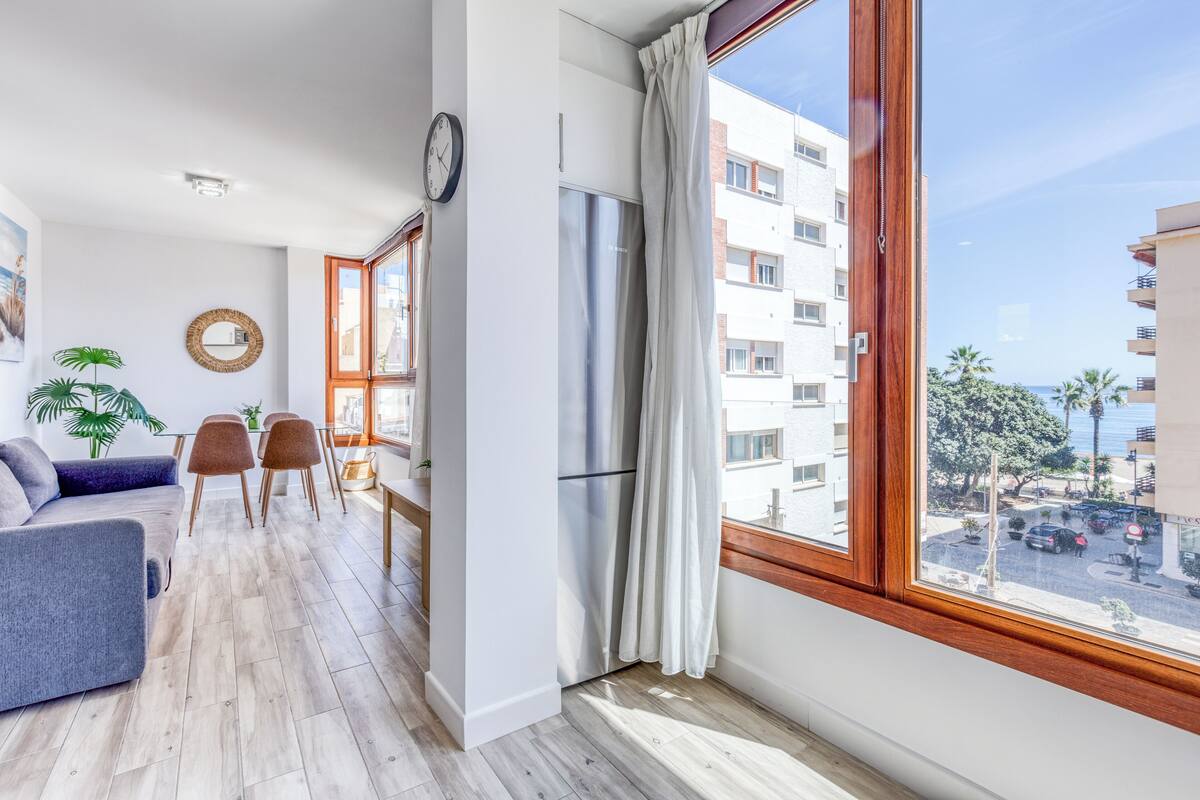 Ein modernes Wohnzimmer mit einer Couch, Sesseln, einem Esstisch und einem großen Fenster mit Vorhängen.