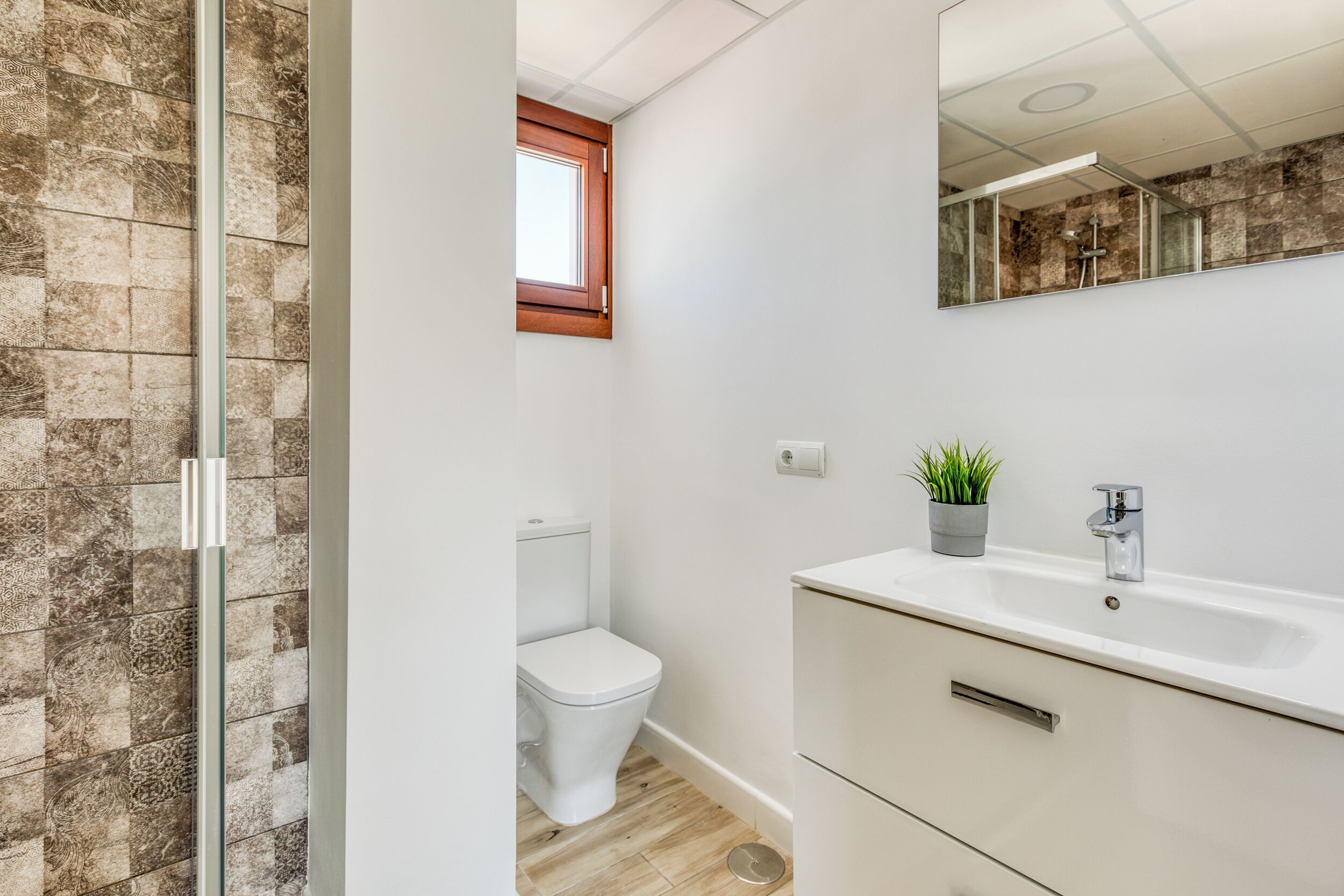Et badeværelse med glasbrusekabine, toilet, vask med spejl og en lille plantekasse.