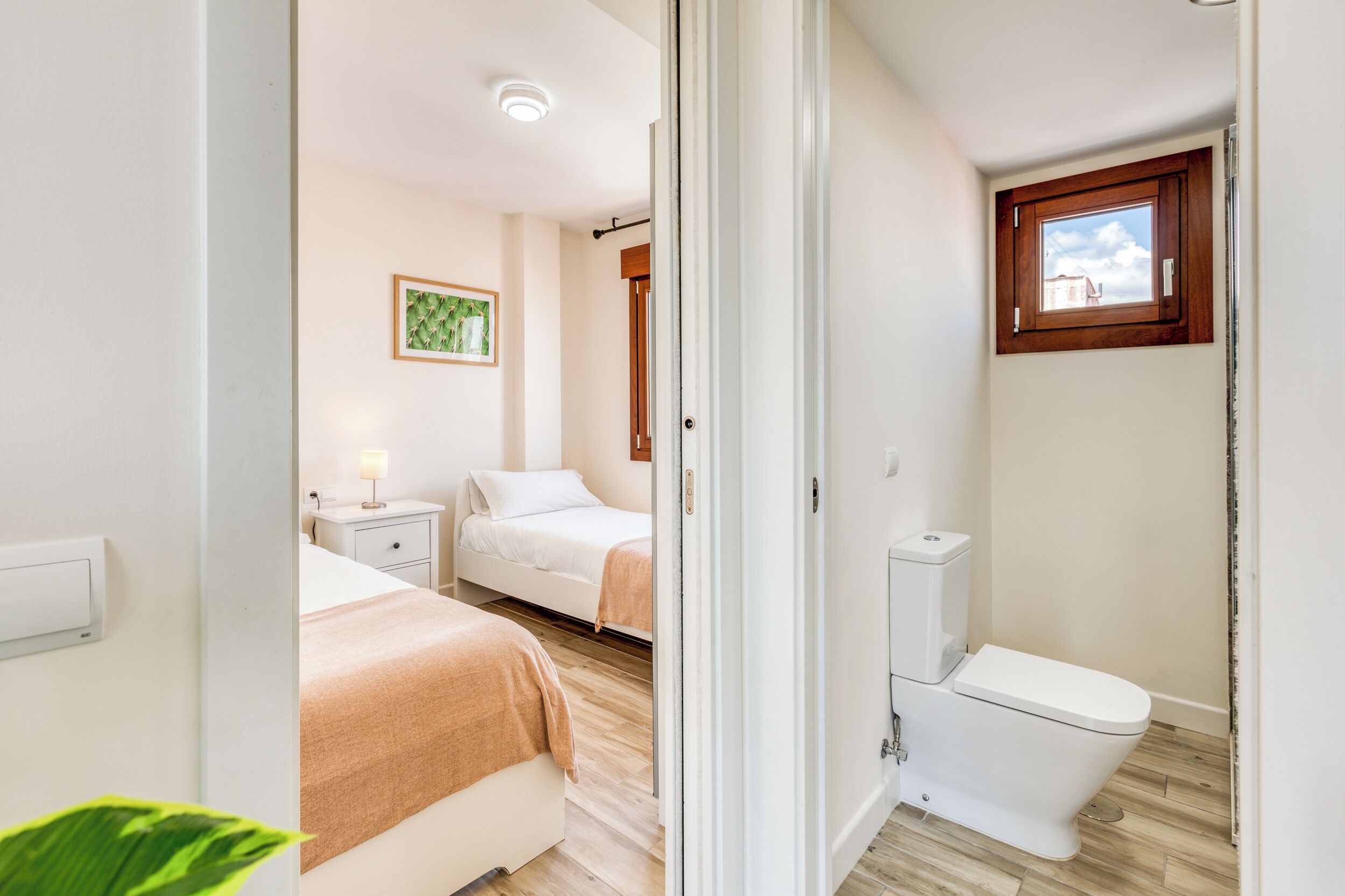 A bedroom with two beds, a bedside table with a lamp, a window with a view, and a white toilet.